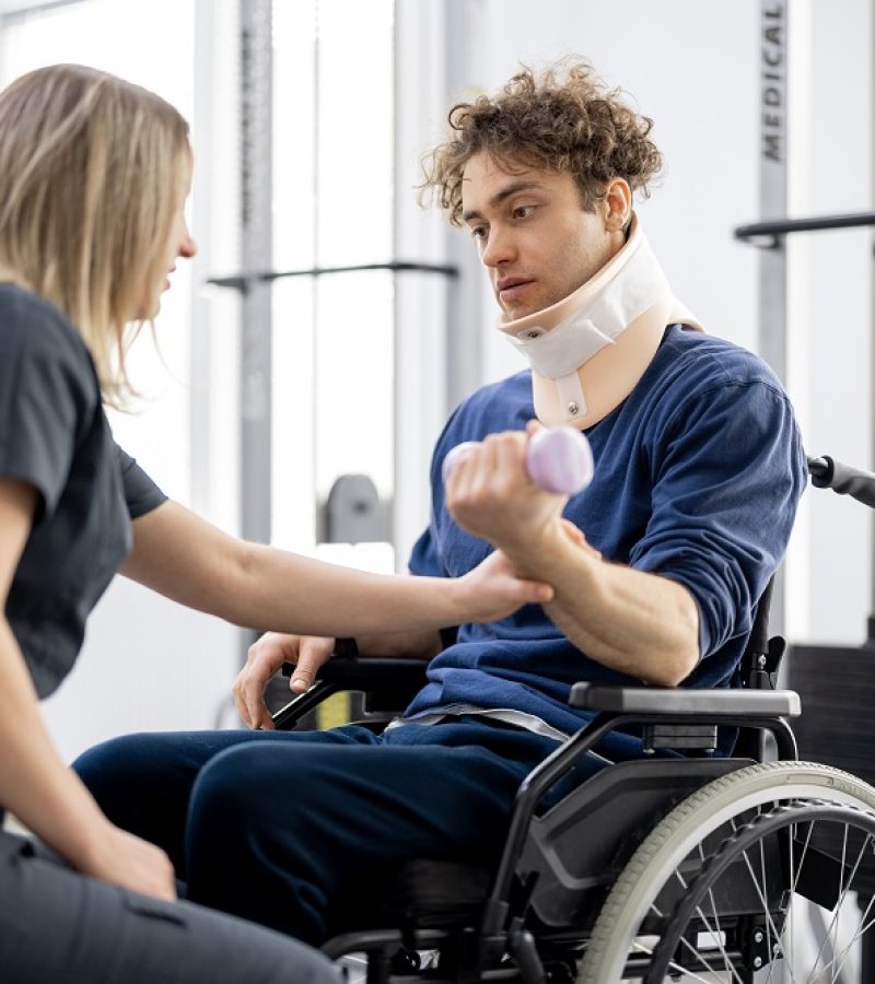 Rehabilitation specialist helps a guy to do exercises for recovery from injury, who is sitting in a wheelchair with a corset around his neck. Concept of physical therapy for people with disabilities